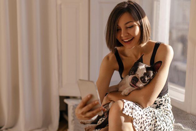 Симпатичная молодая кавказская брюнетка с удовольствием обнимает собаку и делает селфи на телефоне Любовь к животным концепция питомца