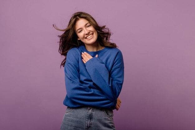 Cute young caucasian brunette woman in blue hoodie smiling while looking at camera on purple background People emotions lifestyle concept