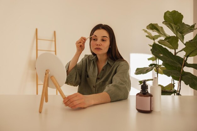 Cute young caucasian woman takes care of her face by applying oil to her eyebrows and eyelashes Brunette girl is sitting at table with mirror Concept of femininity and beauty