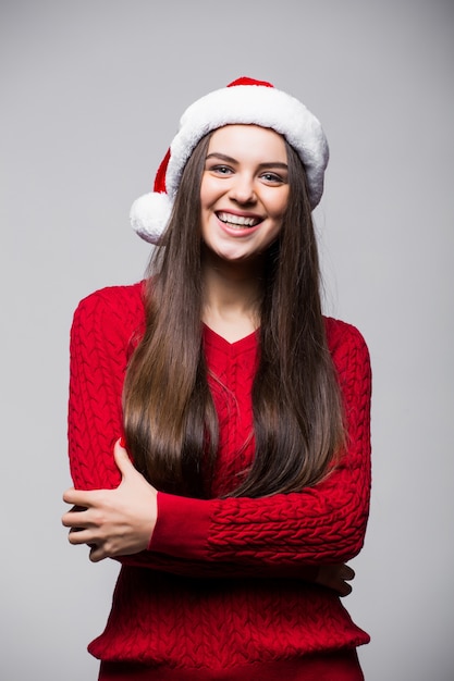 Cute young Caucasian woman wearing Santa hat and gloves posing smiling  against light gray wall. Christmas and New Year concept. Copy space available.