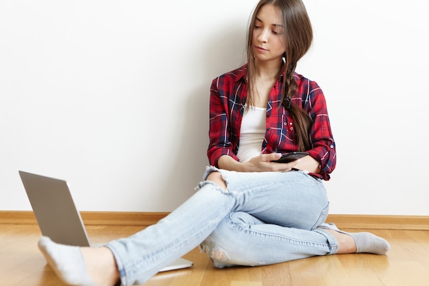 Cute young European female having rest at home wearing stylish clothes holding cell phone