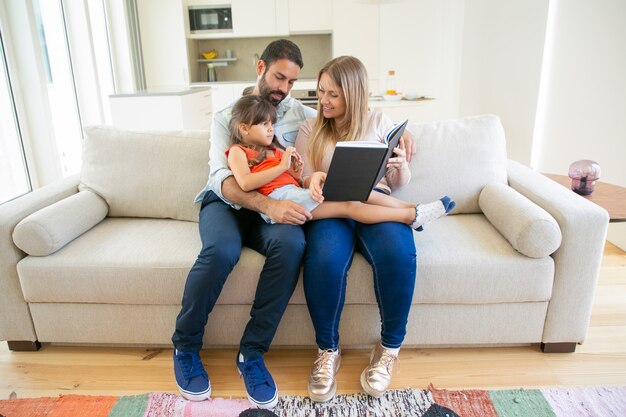 Cute young family couple with little girl sitting on couch in living room and reading book together.