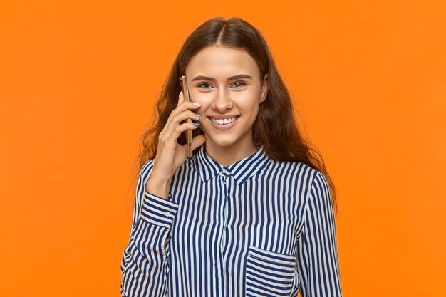 Cute young female holding cell phone at her ear and smiling joyfully, talking to boyfriend