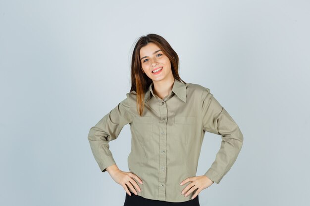 Cute young female holding hands on waist in shirt and looking cheerful. front view.