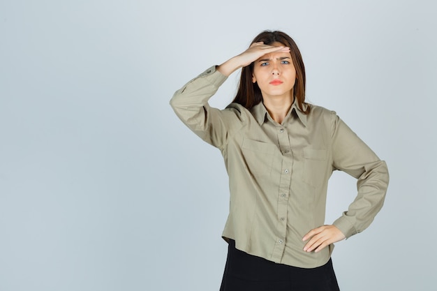 Cute young female in shirt, skirt holding hand over head and looking confused , front view.