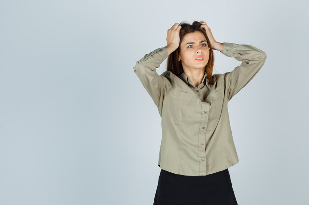 Cute young female in shirt, skirt holding hands on head and looking confused , front view.