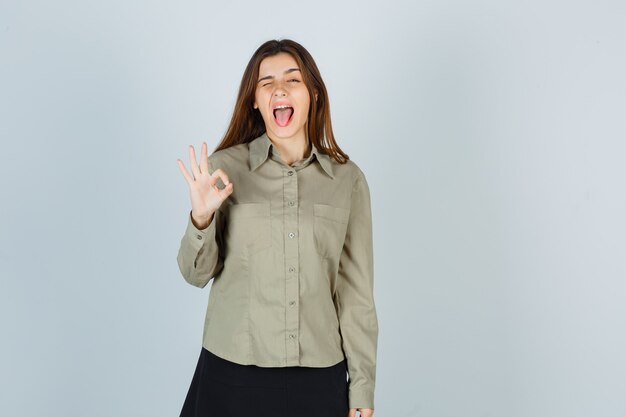 Симпатичная молодая женщина в рубашке, юбке показывает жест ОК, моргая, высунув язык и выглядит сумасшедшим, вид спереди.