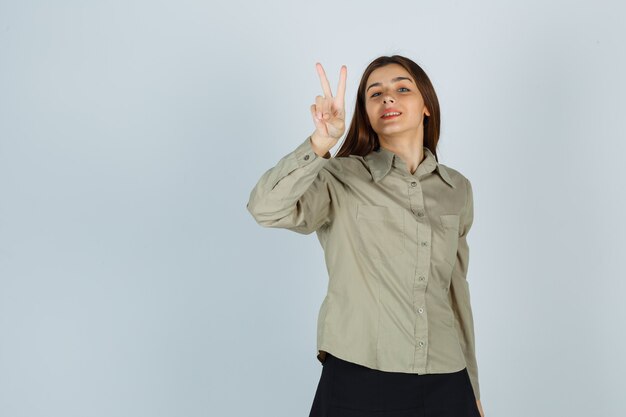 Cute young female in shirt, skirt showing victory gesture and looking merry , front view.