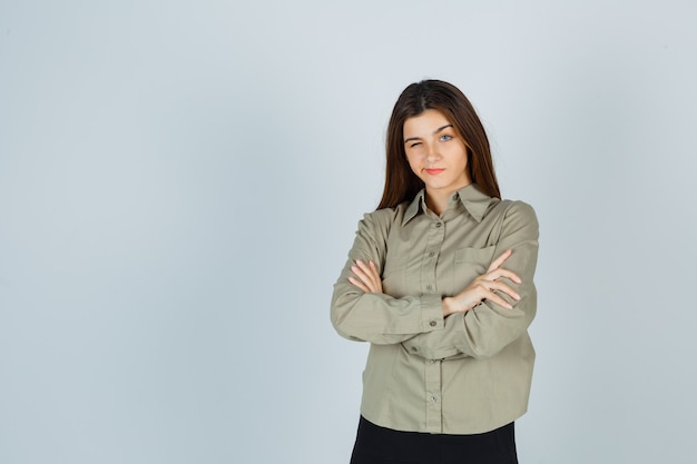 Free photo cute young female in shirt, skirt standing with crossed arms, curving lips, blinking and looking thoughtful , front view.