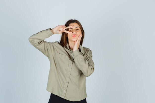 Cute young female showing V-sign near eye while blinking, pouting lips, holding hand on cheek in shirt and looking confident. front view.