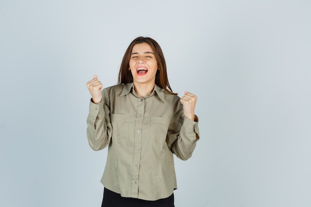 Cute young female showing winner gesture while screaming in shirt and looking blissful , front view.