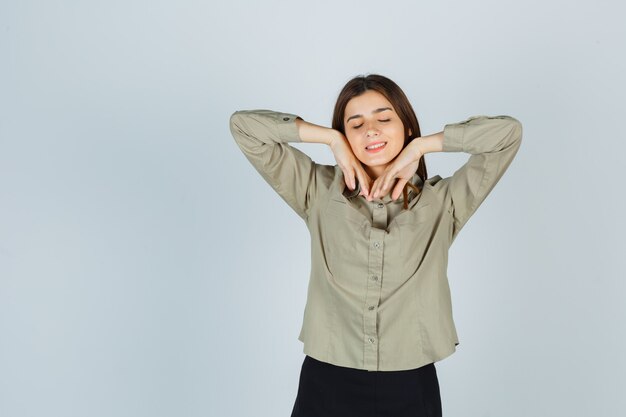 Cute young female stretching elbows while shutting eyes in shirt, skirt and looking delighted. front view.