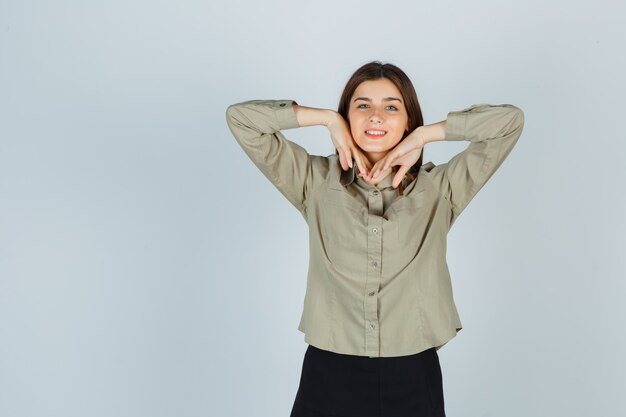 Cute young female stretching elbows while smiling in shirt, skirt and looking merry , front view.