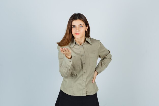 Cute young female stretching hand in questioning gesture in shirt, skirt and looking serious. front view.