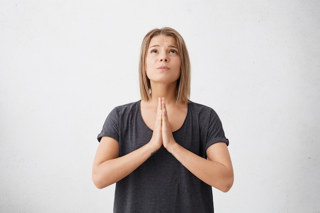 Free photo cute young female wearing casual t-shirt keeping palms pressed together having sorrowful expression looking up begging for good luck in future isolated.