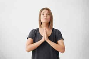 Free photo cute young female wearing casual t-shirt keeping palms pressed together having sorrowful expression looking up begging for good luck in future isolated.