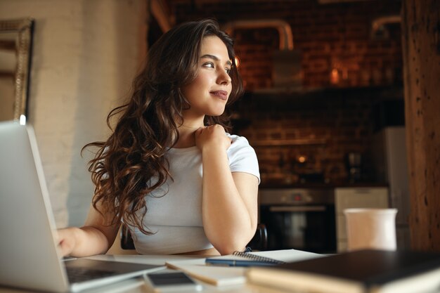 Cute young female with long wavy hair enjoying sunny day using portable computer for distant work, having dreamy look. Charming student girl studying online on laptop at home. Selective focus