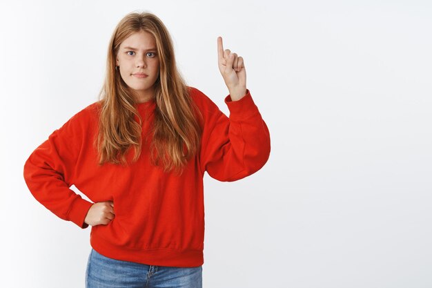 Cute young female with red hair, freckles and blue eyes raising hand pointing up, holding arm on waist