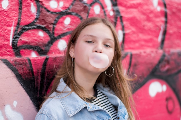 Cute young girl blowing bubble gum against graffiti wall