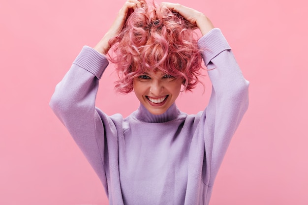 Cute young girl ruffles curly pink hair on isolated wall