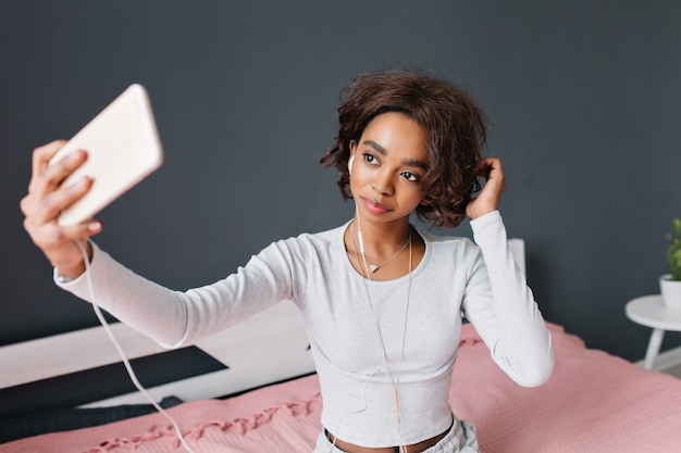 Cute young girl, teen taking selfie, listening to music on bed with pink carpet in room with grey wall. Wearing light gray t-shirt with long sleeves, pendant triangle.