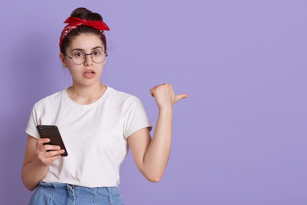 Free photo cute young girl white t-shirt posing against lilac space, holding in hand mobile phone, pointing fingers aside on copy pace