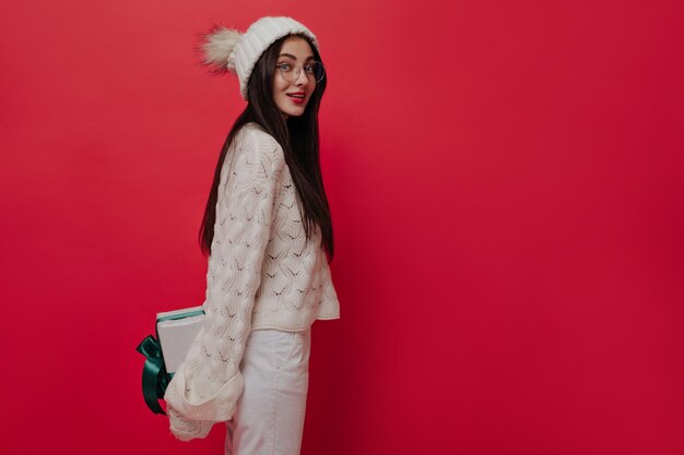 Cute young girl with brunette long hairstyle in light winter outfit bright makeup and glasses holding gift box behind and looking into camera against red background