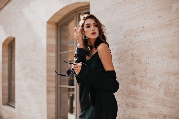 Cute young girl with dark wavy hairstyle and bright makeup, silk dress, black jacket, holding sunglasses in hands and looking away against beige building wall