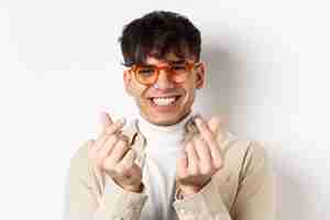 Free photo cute young guy in glasses smiling and showing finger hearts, standing on white background