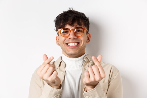 Cute young guy in glasses smiling and showing finger hearts, standing on white background