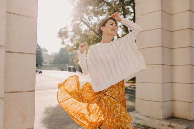 Cute young happy smiling woman in yellow printed dress and knitted white sweater on sunny autumn day