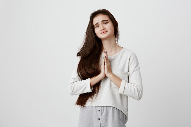 cute young hopeful dark-haired woman wearing casual long sleeved top pressing palms together, feeling worried and desperate while praying for wellness and good health of her parents.