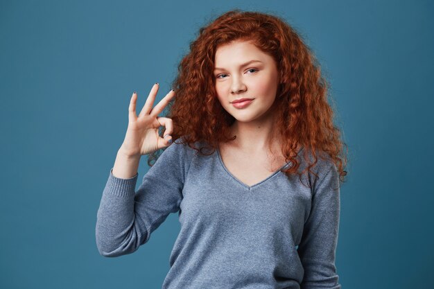 Cute young red haired woman with freckles in grey shirt showing OK gesture with hand, looking with happy and relaxed expression.