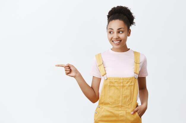 Cute young and tender female sibling with dark skin in yellow trendy overalls, holding hand in pocket, pointing and looking left