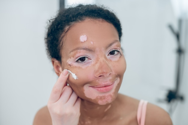 A cute young woman applying moisturizing cream on her face