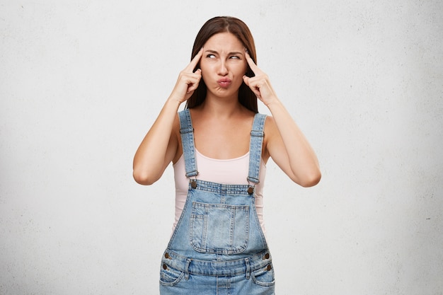 Foto gratuita carina giovane donna in tuta di jeans tenendo le dita sulle tempie e guardando lateralmente con espressione seria concentrata durante la ricerca di una soluzione a problemi personali o problemi sul lavoro