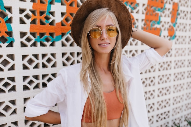 Cute young woman in elegant shirt and hat posing near wall. Outdoor shot of lovable caucasian woman in yellow sunglasses relaxing at resort.