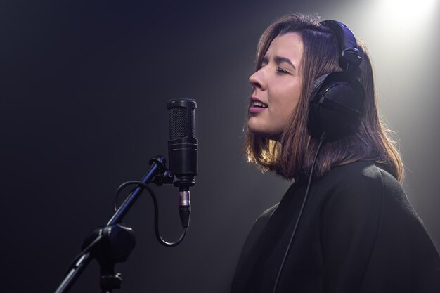 Cute young woman in headphones sings into a microphone in a dark room