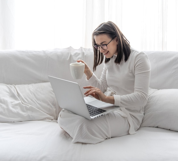 A cute young woman is sitting at home on a white sofa in a white dress in front of a laptop