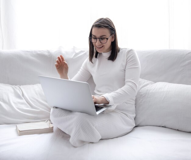 A cute young woman is sitting at home on a white sofa in a white dress and working on a laptop.