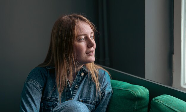 A cute young woman is sitting on the sofa by the window