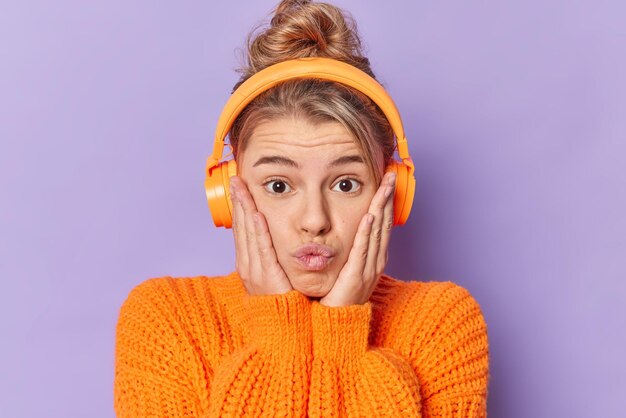 귀여운 젊은 여성이 뺨에 손을 대고 입술을 삐죽 내밀며 보라색 배경에 격리된 니트 스웨터를 입고 무선 헤드폰을 통해 음악을 듣습니다.