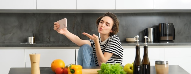 キュートな若い女性ライフスタイルブロガーが 野菜と切り板でキッチンに座って料理をしています