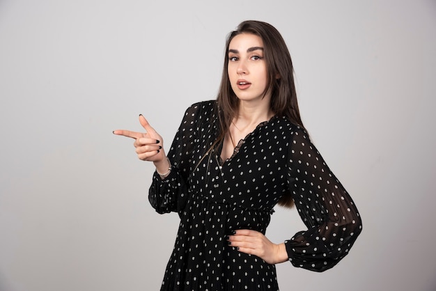 Cute young woman pointing a finger away on a gray wall.