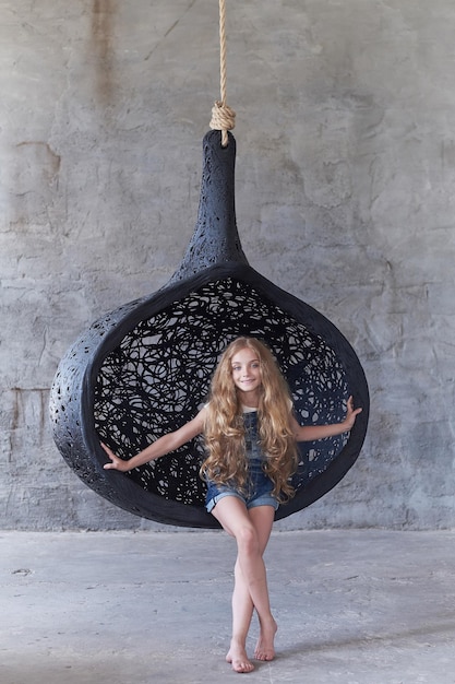 Cute young woman posing near big oval black artistic chair over grey background.