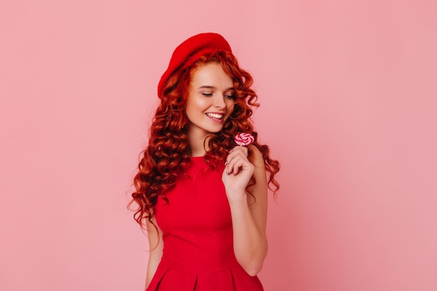 Cute young woman in red dress and felt hat posing with lollipop on pink space.