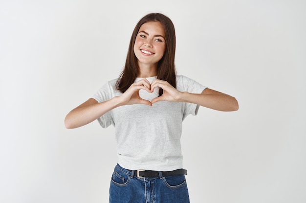 Cute young woman showing heart sign, I love you gesture, wishing happy valentines day, standing over white wall.