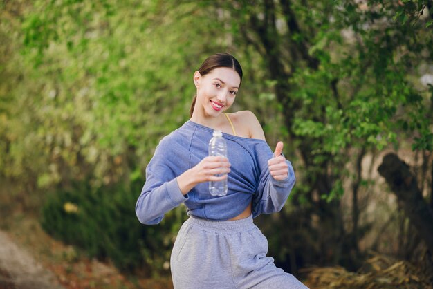 Cute young woman in sport clothes with bottle of water and showing thumb up
