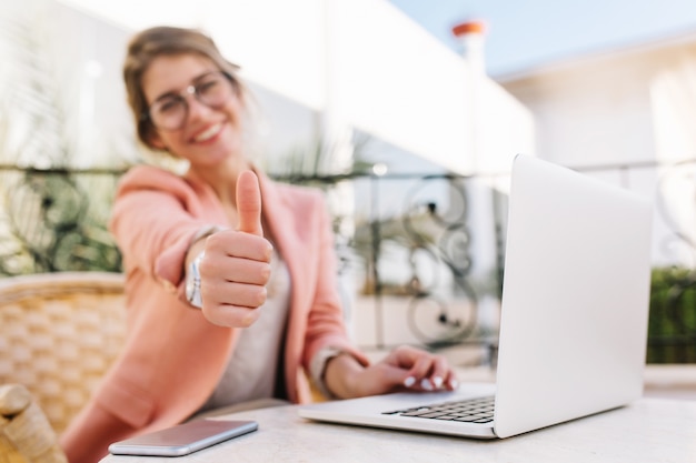 Cute young woman, student, business lady showing thumbs up, well done, sitting in outdoor cafe on terrace with laptop. wearing pink smart clothes. Free Photo