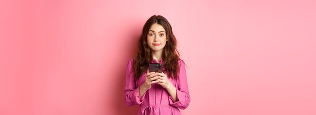 Free photo cute young woman using mobile phone app holding smartphone and smiling at camera standing in dress against pink background